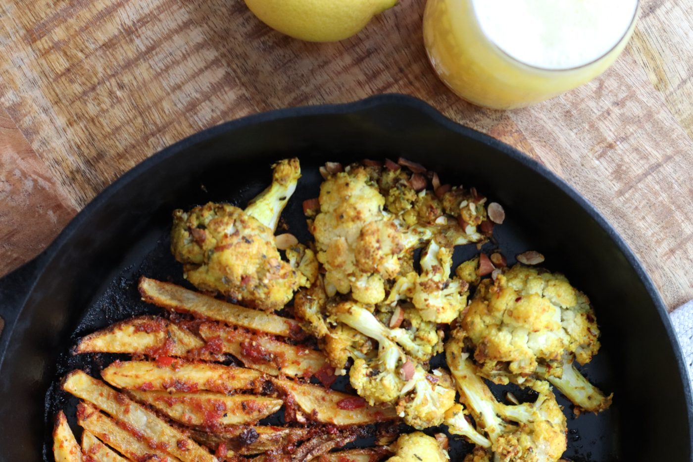 Overhead cast-iron skillet of spiced roasted cauliflower and seasoned fries with citrus juice alongside