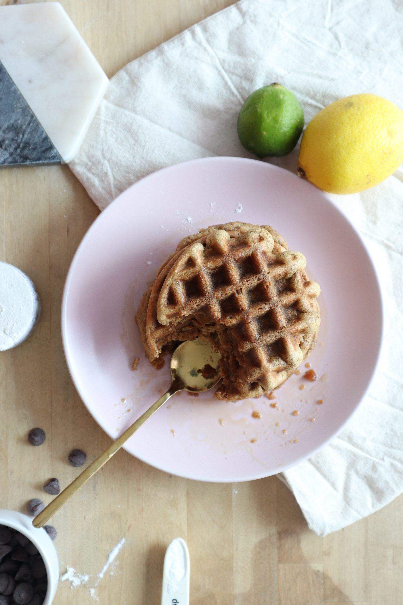Overhead stack of waffles with a bite taken, gold spoon resting in the bite, lime and lemon beside the plate
