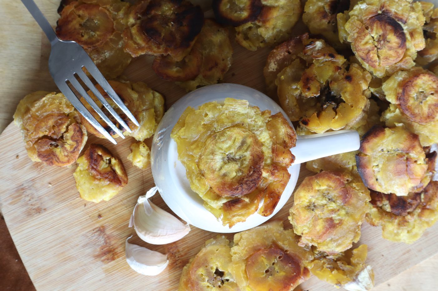 Single tostone served on a small white pinch bowl, surrounded by more tostones, a fork, and garlic cloves on the board